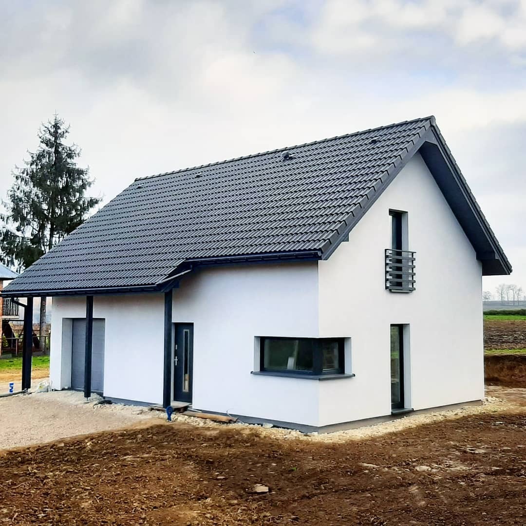 Nowoczesny, parterowy dom z białymi ścianami i ciemnoszarą dachówką karpiówką, widoczny z zewnątrz, z przygotowanym terenem pod ogród.