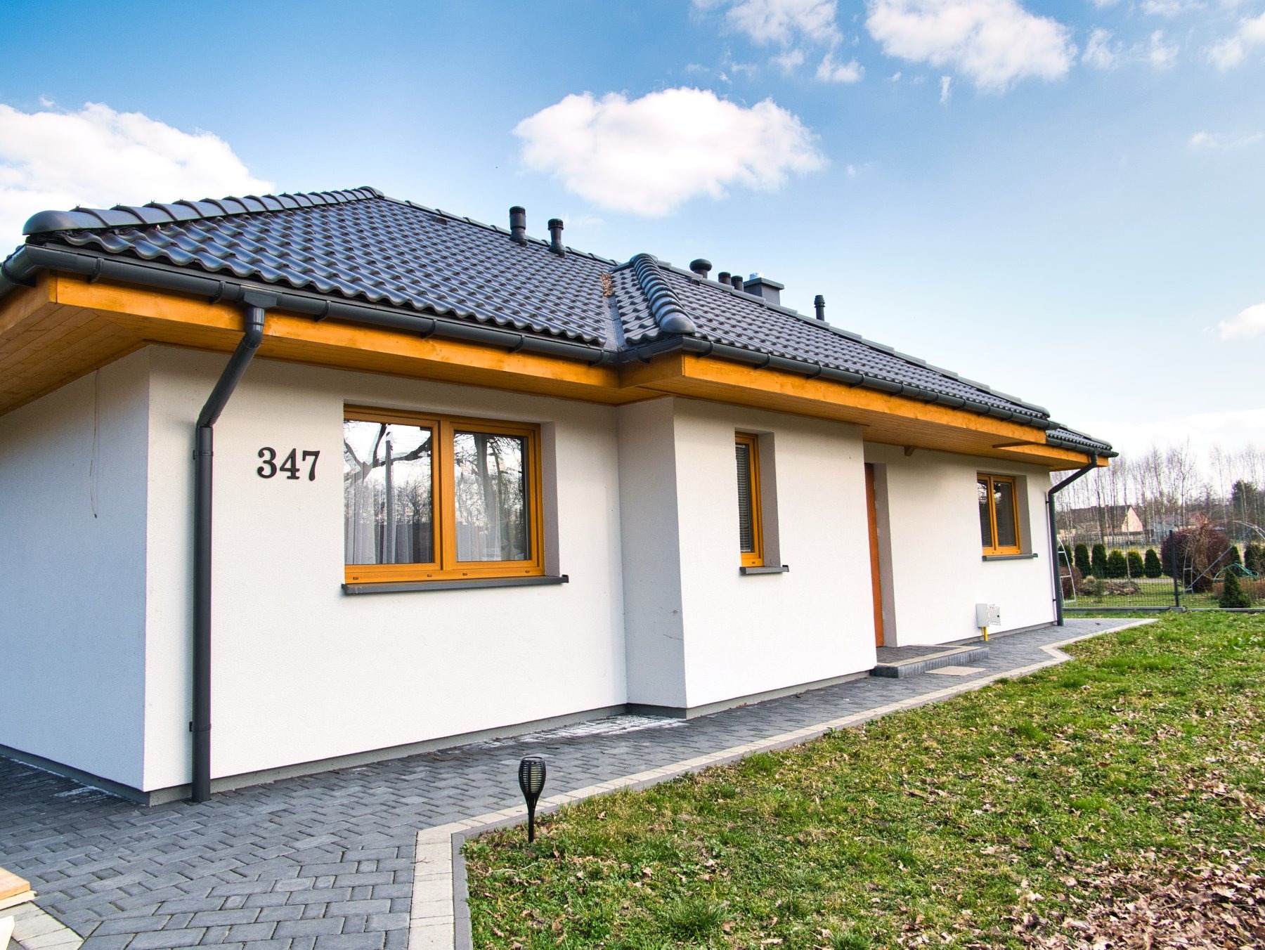 Parterowy dom z białymi ścianami, ciemną dachówką i numerem 347, otoczony trawnikiem i ścieżką z kostki brukowej, widok z zewnątrz w słoneczny dzień.