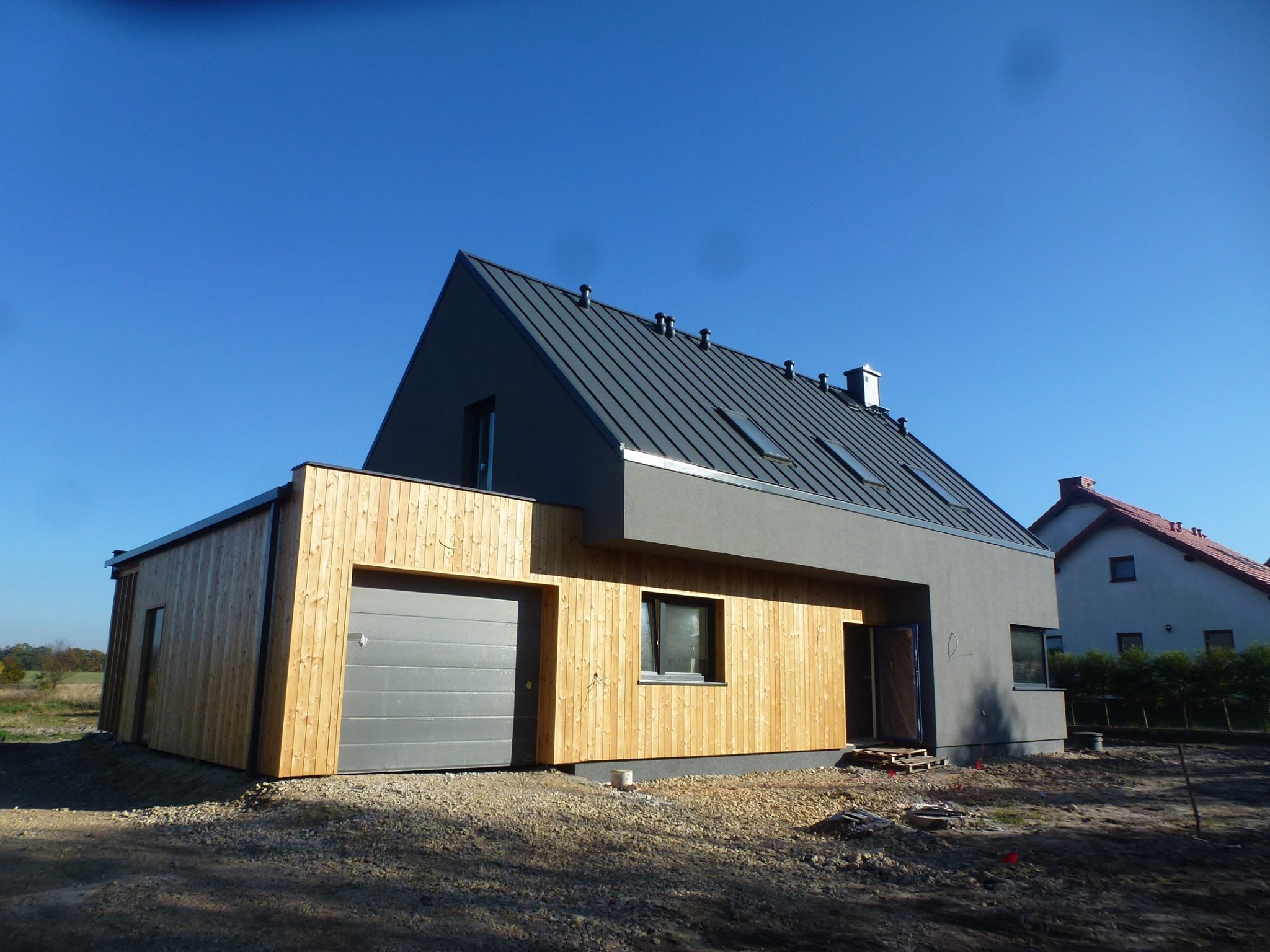 Nowoczesny dom z szarą elewacją i drewnianą fasadą, garaż z szarą bramą segmentową, dach z blachy na rąbek stojący i lukarnami, widok z zewnątrz w słoneczny dzień.