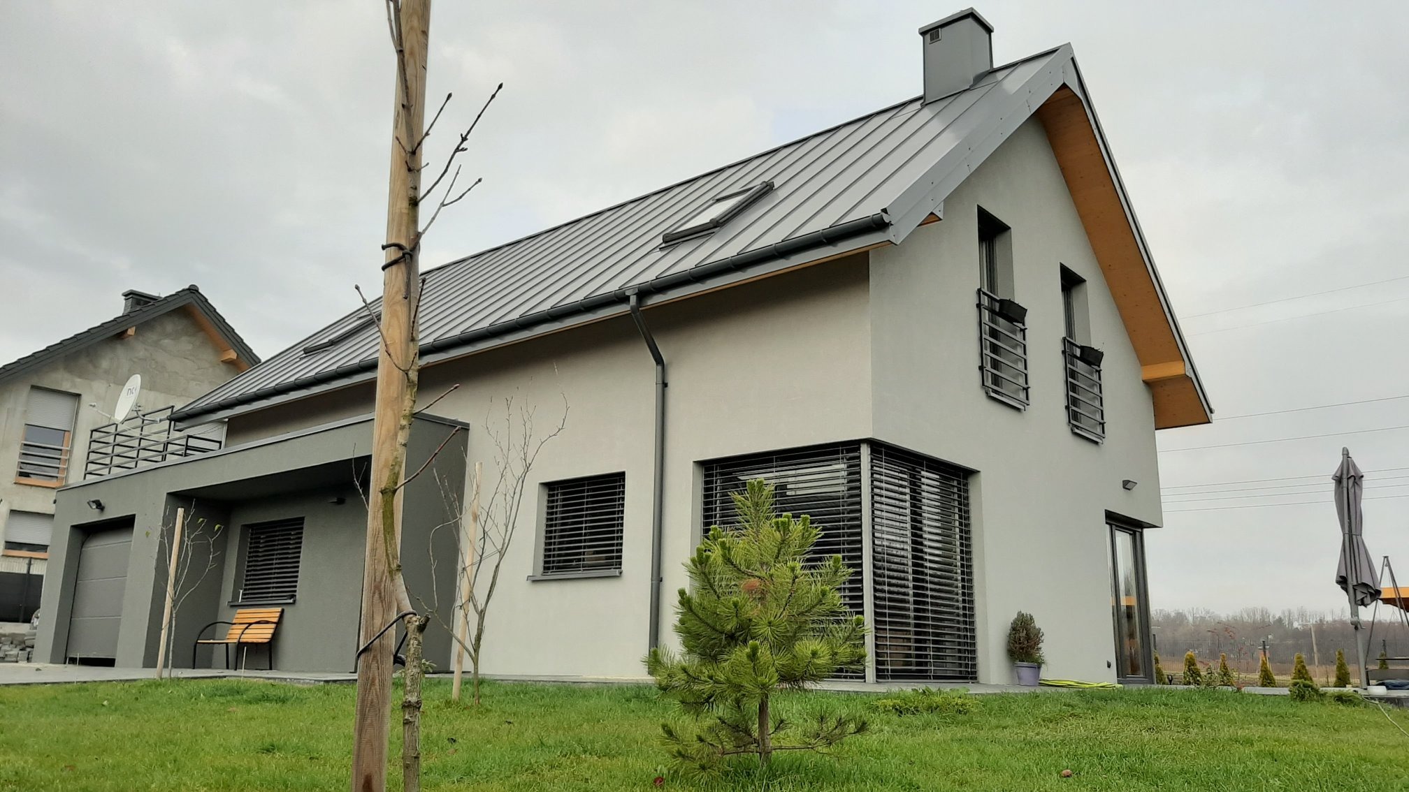 Nowoczesny dom jednorodzinny z szarą blachodachówką i elewacją w kolorze gołębim, z dużymi oknami osłoniętymi żaluzjami zewnętrznymi, na pierwszym planie mała choinka.