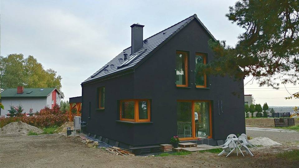 Nowoczesny dom z ciemną elewacją i drewnianymi ramami okiennymi, widoczny na etapie wykończenia terenu, z białymi krzesłami ogrodowymi na pierwszym planie.