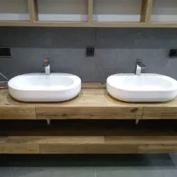 Dwie białe, owalne umywalki nablatowe z chromowanymi bateriami, osadzone na drewnianym blacie z szufladami, na tle szarej ściany z gniazdkami elektrycznymi i wiszącymi szafkami bez frontów.