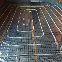 Rozłożone rury ogrzewania podłogowego na folii z siatką, mocowane plastikowymi klipsami w nowo budowanym pomieszczeniu z widocznymi cegłami.