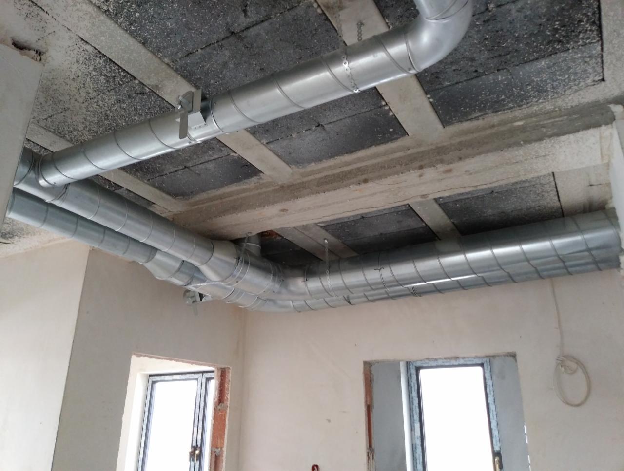 Instalacja kanałów wentylacyjnych z metalu pod sufitem w surowym wnętrzu, widoczne okna i betonowa konstrukcja sufitu.