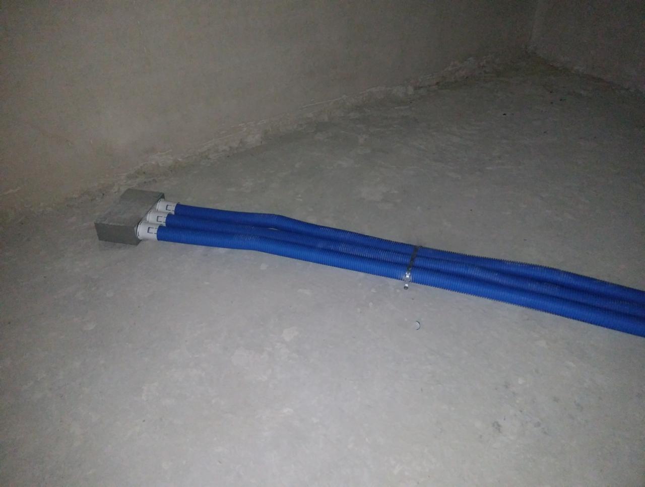 Trzy niebieskie, karbowane rury wychodzące ze ściany, zabezpieczone opaską zaciskową, na surowym, betonowym podłożu w narożniku pomieszczenia.