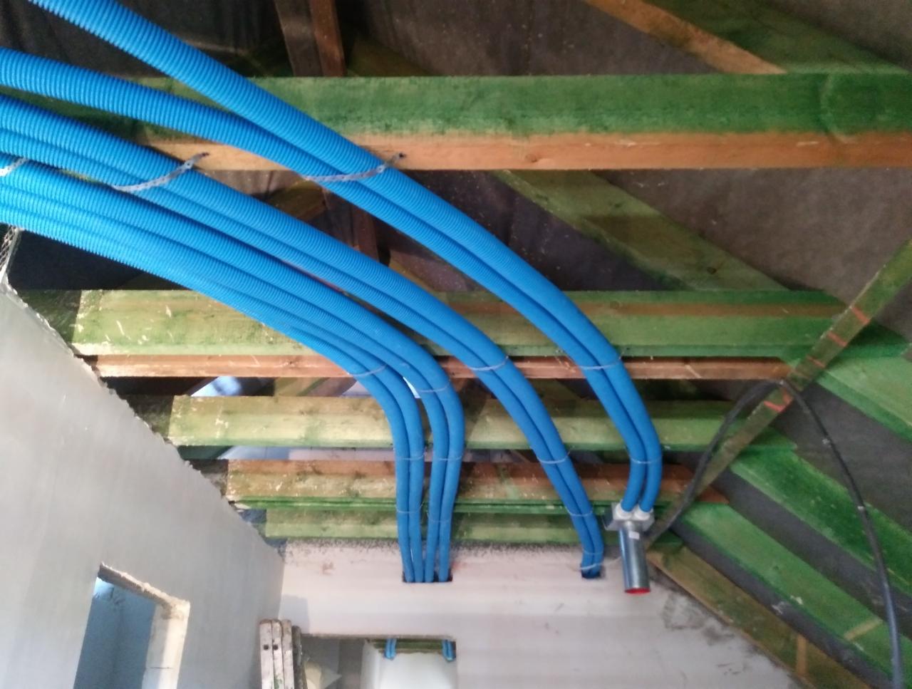 Niebieskie, karbowane rury wentylacyjne poprowadzone pod drewnianą konstrukcją dachu, widoczne elementy instalacji i surowy stan wykończenia wnętrza.