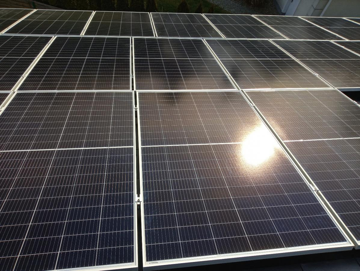 Rząd paneli słonecznych na dachu, z odbiciem słońca tworzącym jasną plamę na powierzchni jednego z paneli.