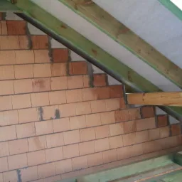 Wnętrze domu w trakcie budowy: ściana z cegły ceramicznej, drewniana konstrukcja dachu z membraną. Widok z dołu na poddasze, surowy stan.