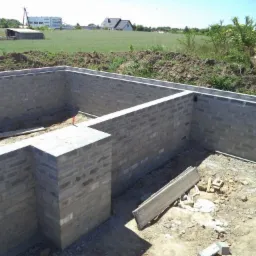 Fundamenty domu z bloczków betonowych, widok z góry. W tle zielone pole i zabudowania. Elementy szalunku widoczne na krawędziach ścian fundamentowych.