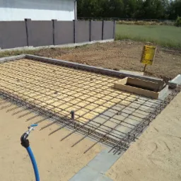 Zbrojenie fundamentu domu jednorodzinnego, widok z góry. Siatka z prętów stalowych na podłożu piaskowym, przygotowana do zalania betonem. Kran z niebieskim wężem w rogu.