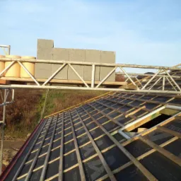 Wznoszenie więźby dachowej z deskowaniem i izolacją. Na rusztowaniu pustaki i kominy. Ujęcie z góry, widoczny krajobraz w tle.