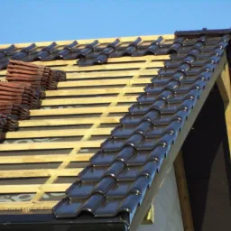 Układanie brązowej dachówki na drewnianej konstrukcji dachu w słoneczny dzień. Częściowo pokryty dach z widocznymi stosami dachówek do ułożenia.