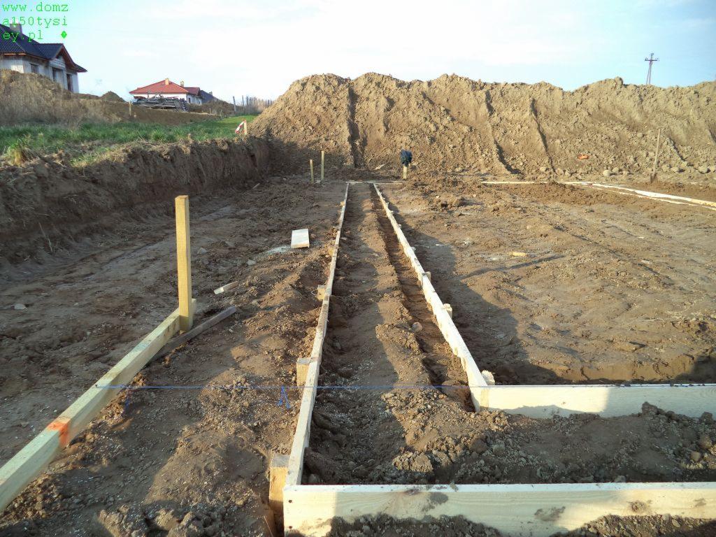 Przygotowanie szalunków pod fundamenty na placu budowy, widoczne drewniane konstrukcje i wykop ziemny. W tle hałda ziemi i domy w oddali.