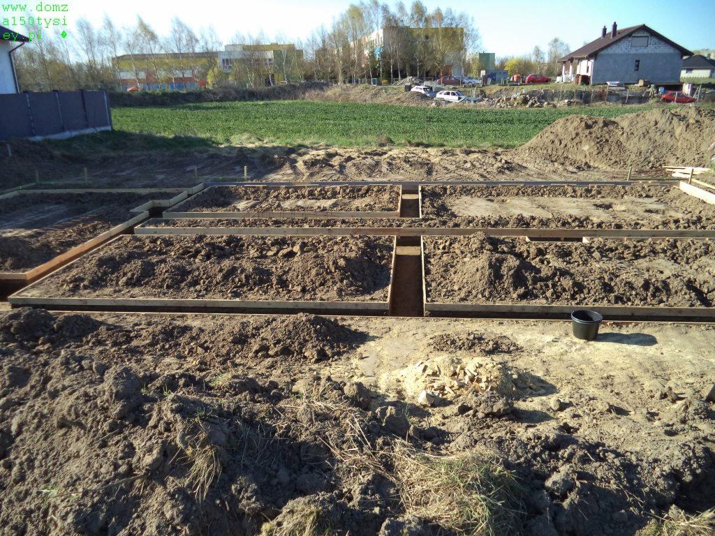 Przygotowanie terenu pod fundamenty: wykopane rowy z drewnianym szalunkiem, ziemia, widok na okolicę z budynkami i zielenią w tle. Czarny kubeł w prawym dolnym rogu.