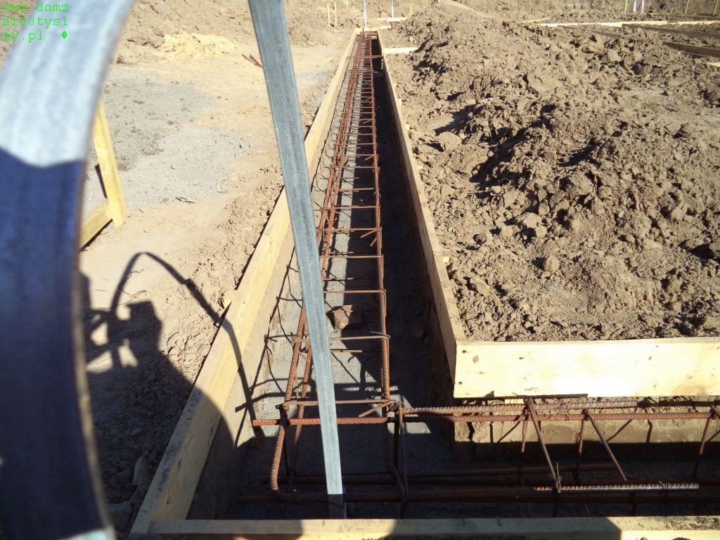 Zbrojenie fundamentów w drewnianym szalunku, widok z góry. Stalowe pręty połączone w siatkę, ziemia w tle. Przygotowanie do wylewania betonu.