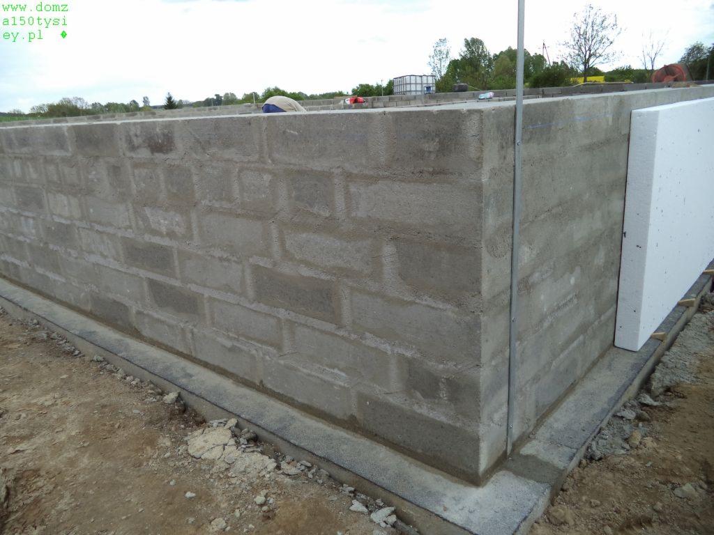 Surowe fundamenty domu z betonu komórkowego, częściowo ocieplone styropianem, widoczne zbrojenia pionowe, teren budowy.