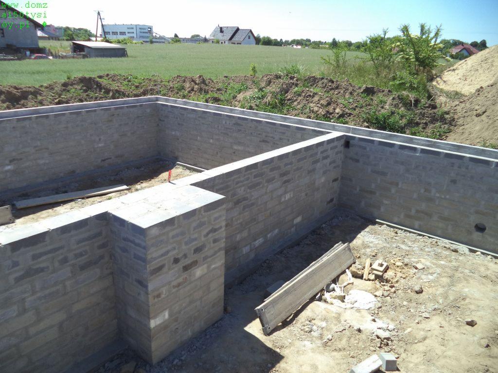 Fundamenty domu z bloczków betonowych, widok z góry. W tle zielone pole i zabudowania. Elementy szalunku widoczne na krawędziach ścian fundamentowych.