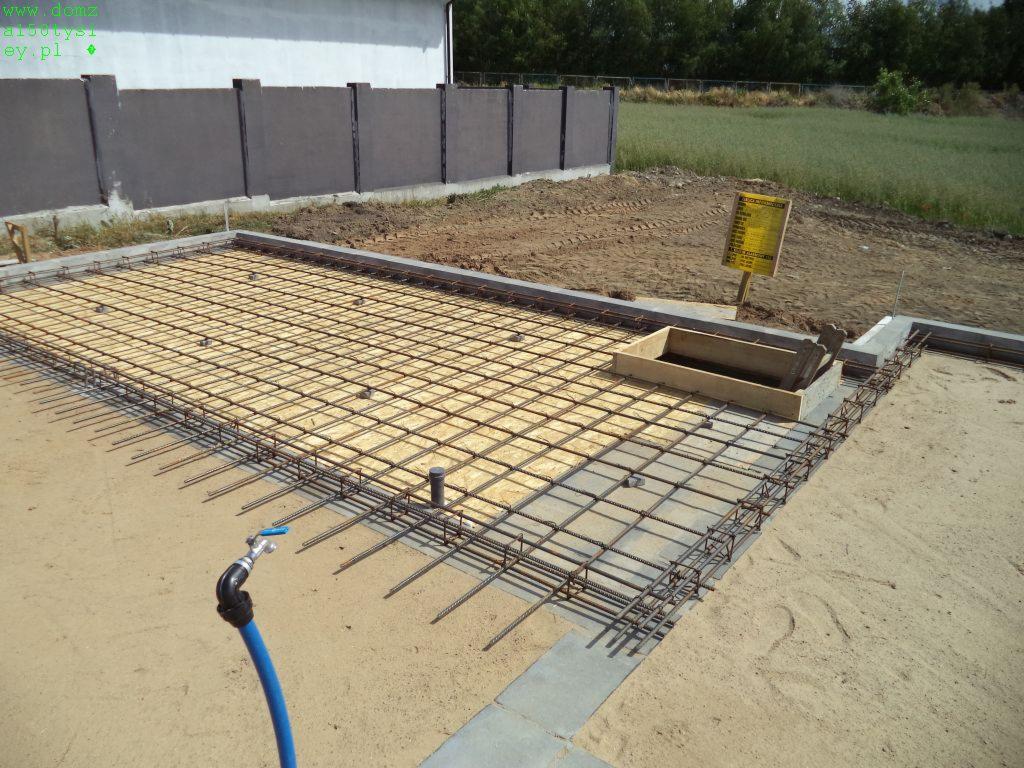 Zbrojenie fundamentu domu jednorodzinnego, widok z góry. Siatka z prętów stalowych na podłożu piaskowym, przygotowana do zalania betonem. Kran z niebieskim wężem w rogu.
