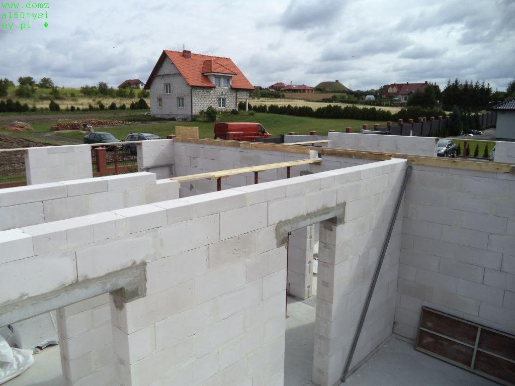 Budowa domu z białych bloczków: ściany w trakcie wznoszenia, widoczne drewniane elementy konstrukcyjne, w tle dom z czerwoną dachówką i zielone otoczenie.