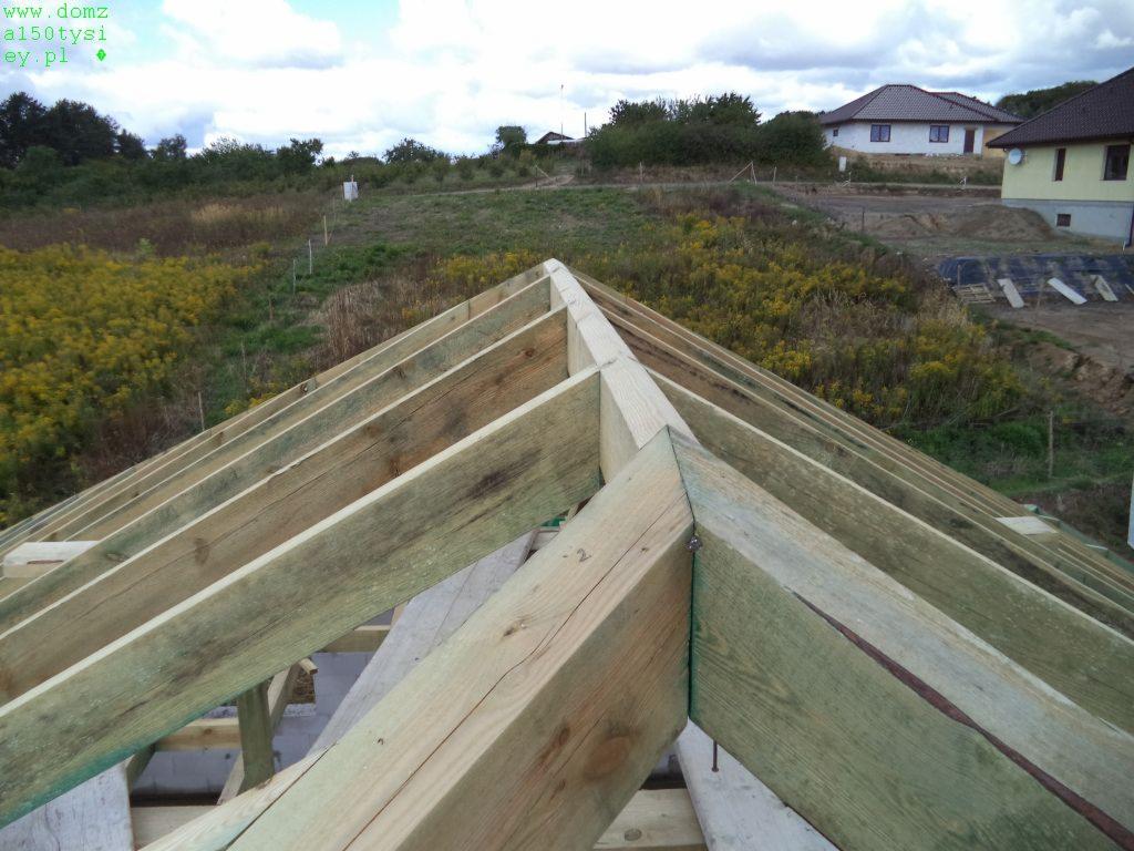 Drewniana konstrukcja dachu w trakcie budowy, widok z góry na krokwie i elementy więźby. W tle krajobraz z budynkami w oddali i zachmurzone niebo.