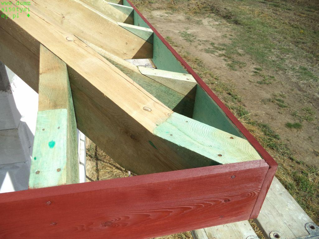 Fragment drewnianej konstrukcji dachu w trakcie budowy, zaimpregnowane elementy w odcieniach zieleni i czerwieni, widok z góry na tle ziemi.