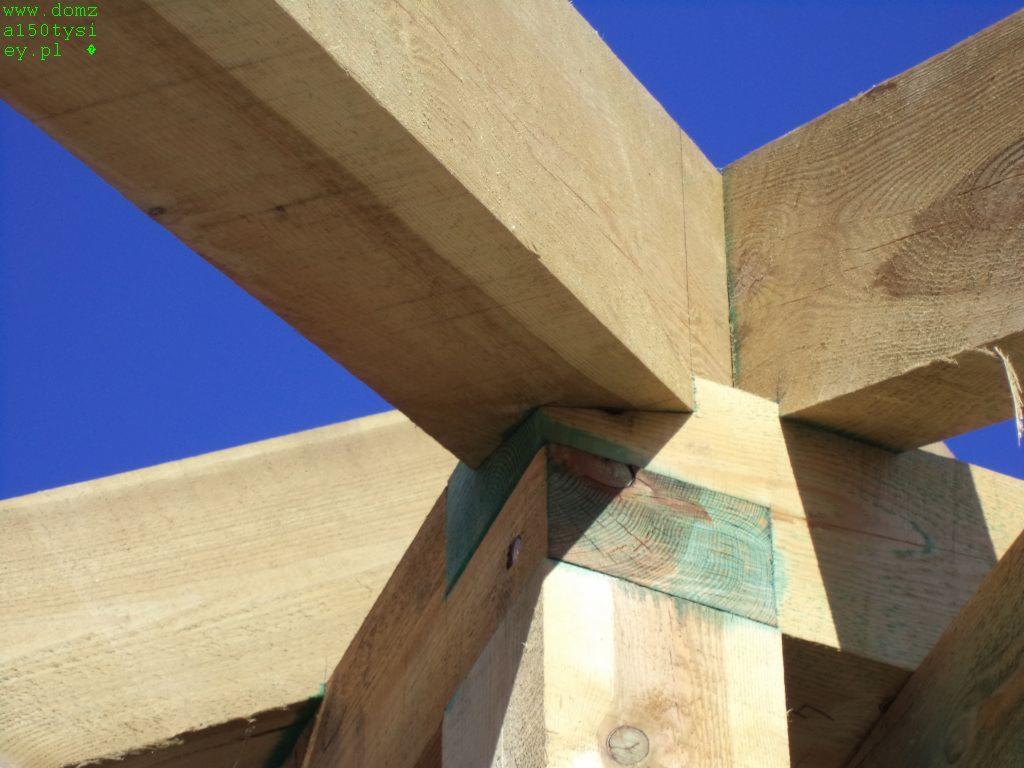 Fragment drewnianej konstrukcji dachu w trakcie budowy, z widocznymi elementami złączeń i impregnacji, na tle błękitnego nieba.