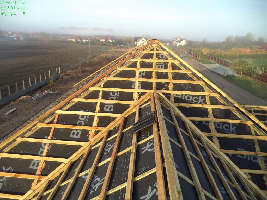 Drewniana konstrukcja dachu z czarną membraną, widok z góry. W tle zabudowania i pole w porannej mgle. Niewielki fragment dachówki na konstrukcji.