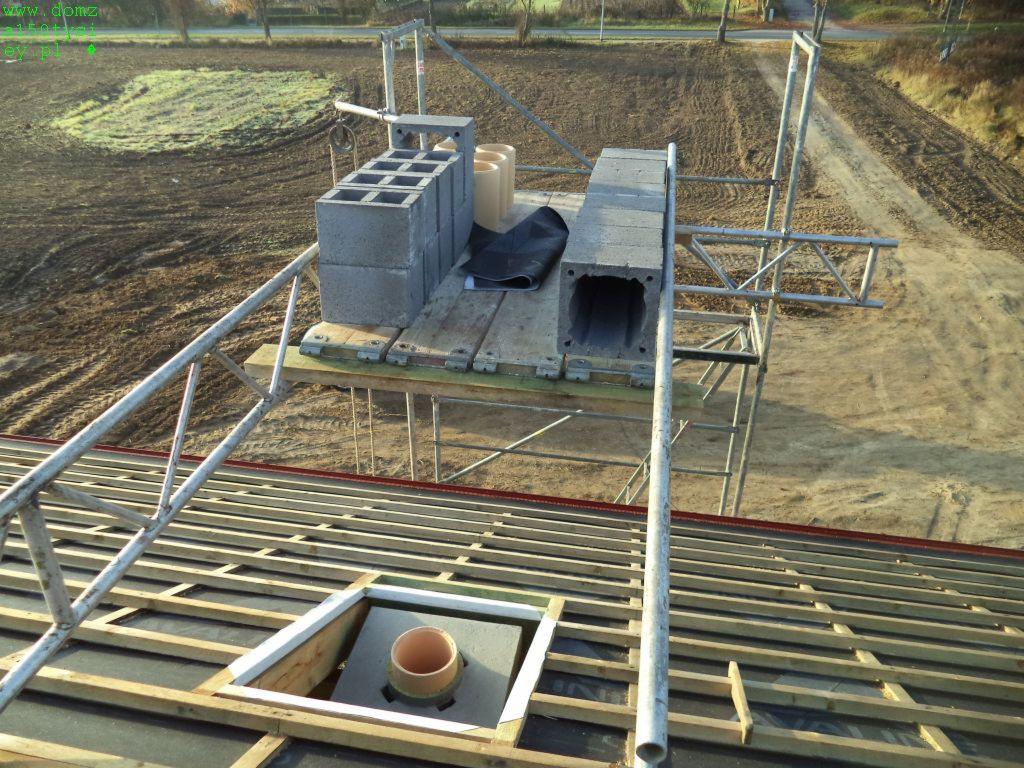 Wznoszenie komina z prefabrykatów na więźbie dachowej z deskowaniem i papą, widok z rusztowania, elementy murowane komina ułożone na podeście roboczym.