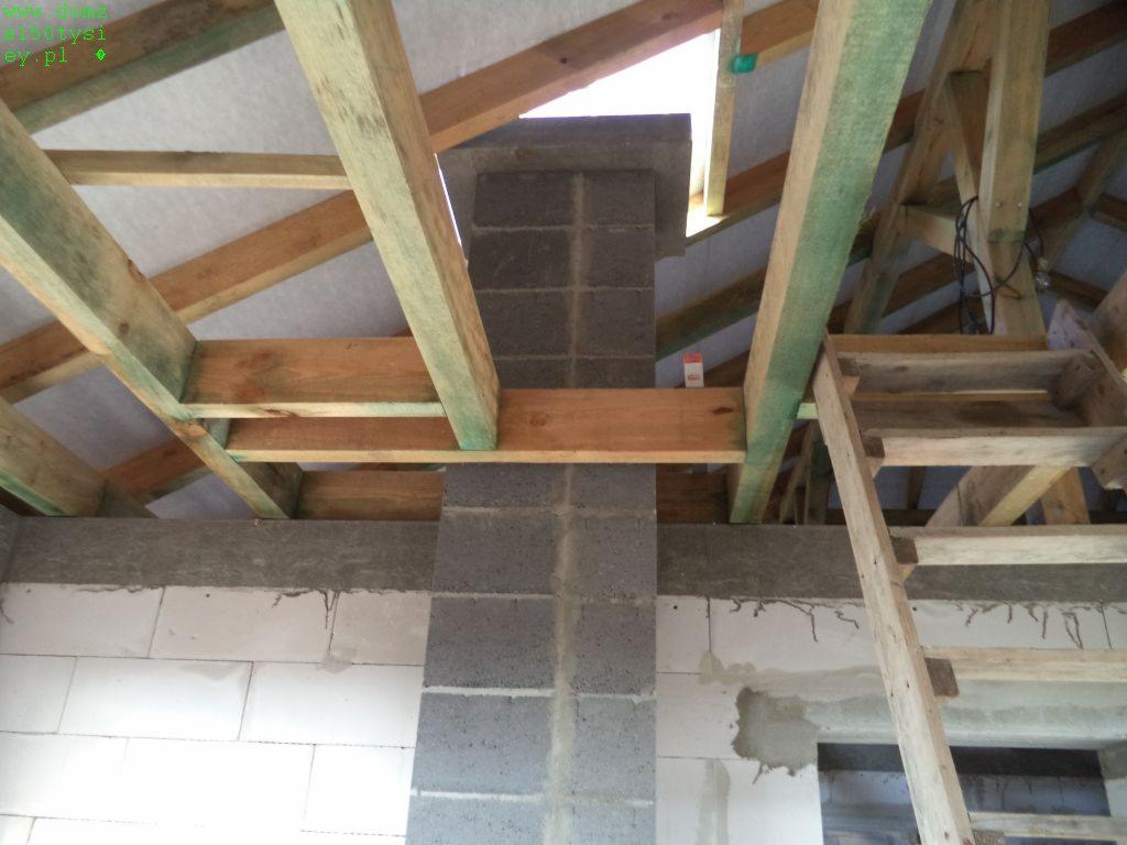 Wnętrze budowy: surowy stan z drewnianą konstrukcją dachu, kominem z bloczków betonowych i fragmentem ściany z betonu komórkowego. Widoczna drabina.