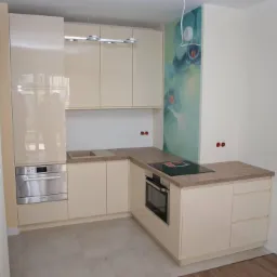 Jasna kuchnia w kształcie litery L z błyszczącymi frontami, płyta indukcyjna i piekarnik w zabudowie, dekoracyjny panel ścienny z motywem kwiatowym, minimalistyczne oświetlenie sufitowe.