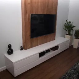 Biała szafka RTV z szufladami i otwartymi półkami, na której stoi telewizor umieszczony na panelu z drewnopodobnym dekorem. Obok szafki dekoracyjne figurki i wysokie donice z roślinami.