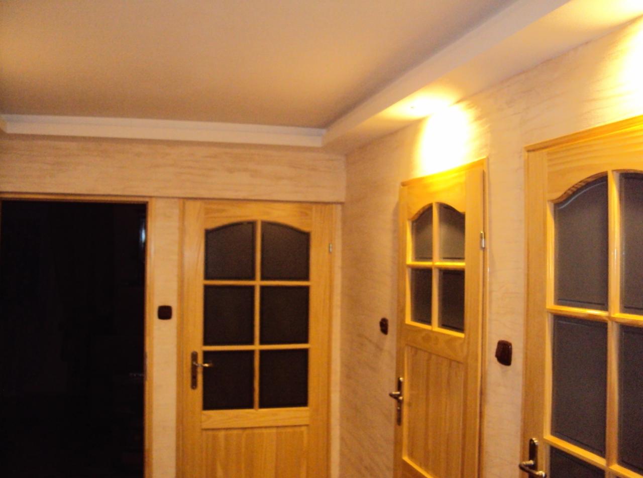 Wykończone wnętrze: drewniane drzwi z szybami, ściany z dekoracyjną fakturą i listwa sufitowa. Ciepłe oświetlenie podkreśla detale wykończenia.