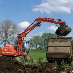 Pomarańczowa koparka Atlas ładuje ziemię do przyczepy na tle błękitnego nieba z chmurami, w otoczeniu zieleni. Widoczny operator w kabinie maszyny.