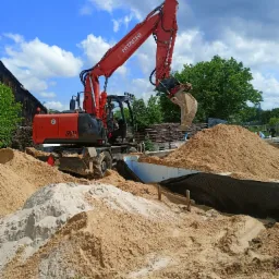 Czerwona koparka Hitachi Zaxis 140W przy wykopie fundamentowym, z hałdami piasku i widocznym zbrojeniem, na tle błękitnego nieba z chmurami.