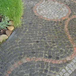 Nawierzchnia z ciemnych, mokrych kamieni ułożonych w łukowaty wzór z akcentami jaśniejszych i czerwonych kamieni, obok rabata z roślinami i kamieniami.