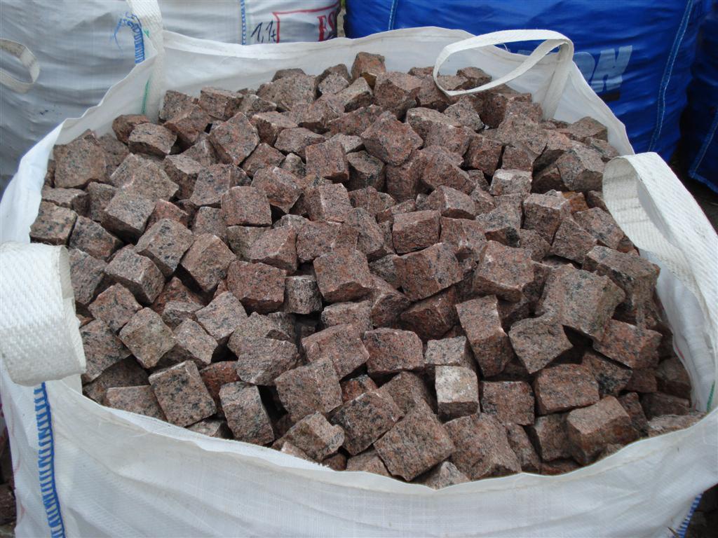 Czerwono-brązowa kostka granitowa, o nieregularnych kształtach, nasypana do worka typu big-bag z białymi uchwytami, widoczne inne worki w tle.
