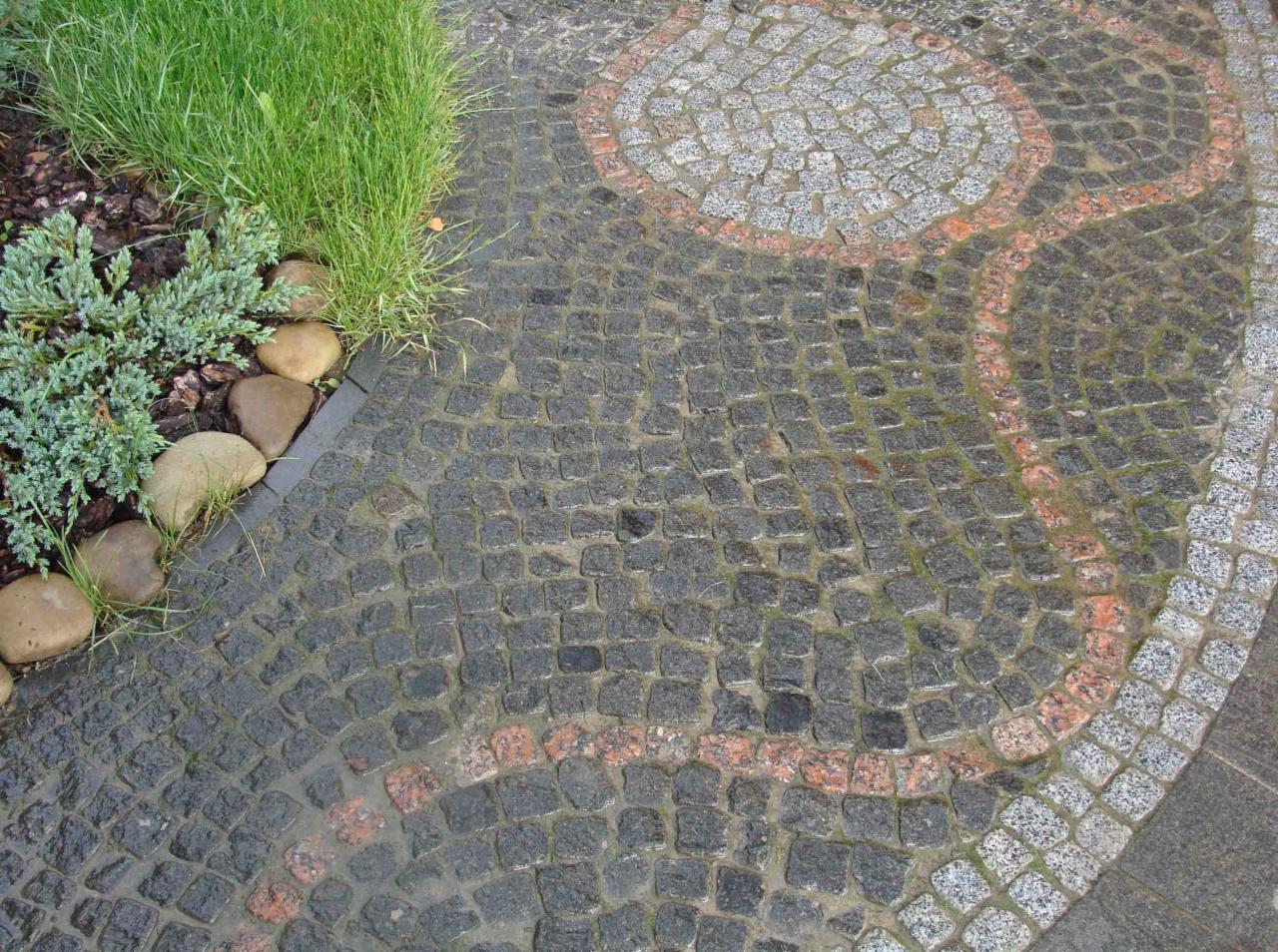 Nawierzchnia z ciemnych, mokrych kamieni ułożonych w łukowaty wzór z akcentami jaśniejszych i czerwonych kamieni, obok rabata z roślinami i kamieniami.