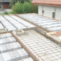 Zbrojenie i betonowe pustaki na stropie budynku w trakcie budowy, widok z góry. Elementy drewniane szalunku i budynki w tle.