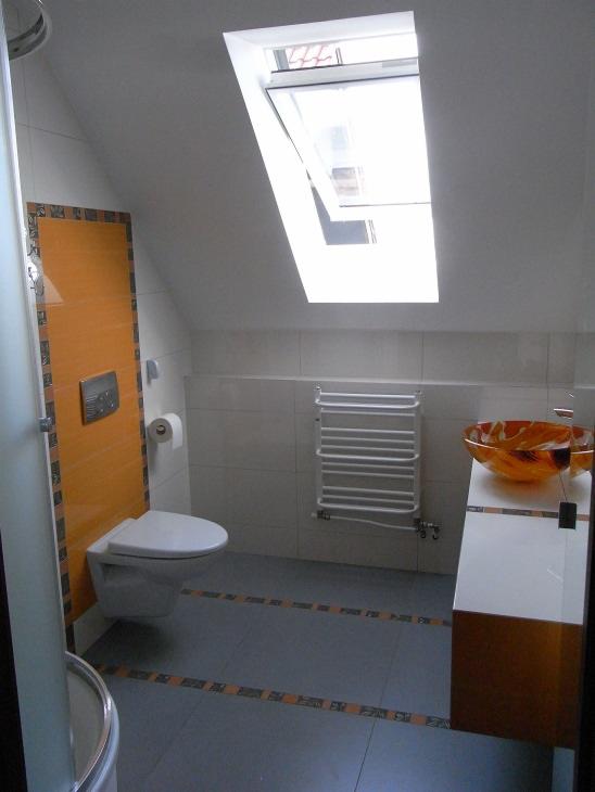 Jasna łazienka na poddaszu ze skośnym sufitem i oknem dachowym. Umywalka w odcieniach pomarańczy, biała toaleta i grzejnik drabinkowy. Dekoracyjne płytki podłogowe.