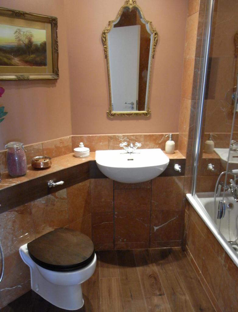 Wykończona łazienka w ciepłych tonacjach z marmurowymi płytkami, drewnianą podłogą i ozdobnym lustrem nad umywalką. Widoczny fragment wanny z kabiną.