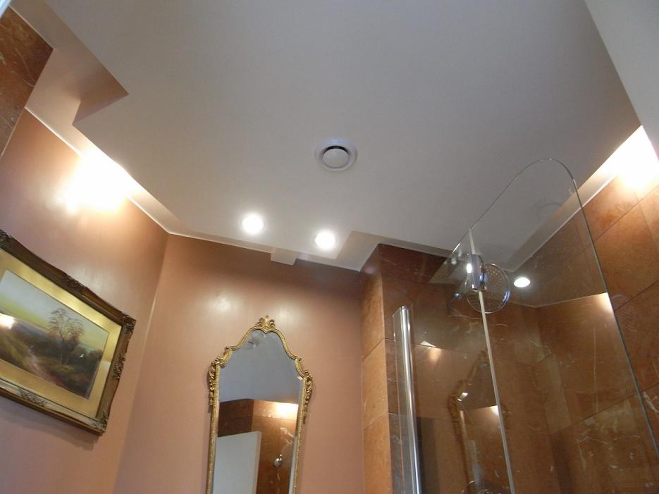 Wykończona łazienka z beżowymi ścianami, dekoracyjnym lustrem i kabiną prysznicową ze szklaną ścianką. Sufit podwieszany z oświetleniem punktowym i kratką wentylacyjną.