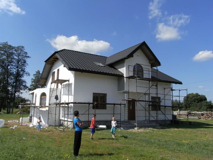 Dom jednorodzinny w trakcie ocieplania elewacji, z widocznymi rusztowaniami i fragmentami styropianu. W tle błękitne niebo z chmurami i osoby w ogrodzie.