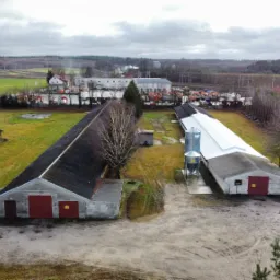 Widok z lotu ptaka na kompleks budynków gospodarczych z ciemnymi dachami i czerwonymi bramami, uzupełnionych o metalowe silosy, na tle rozległych pól i lasów pod pochmurnym niebem.