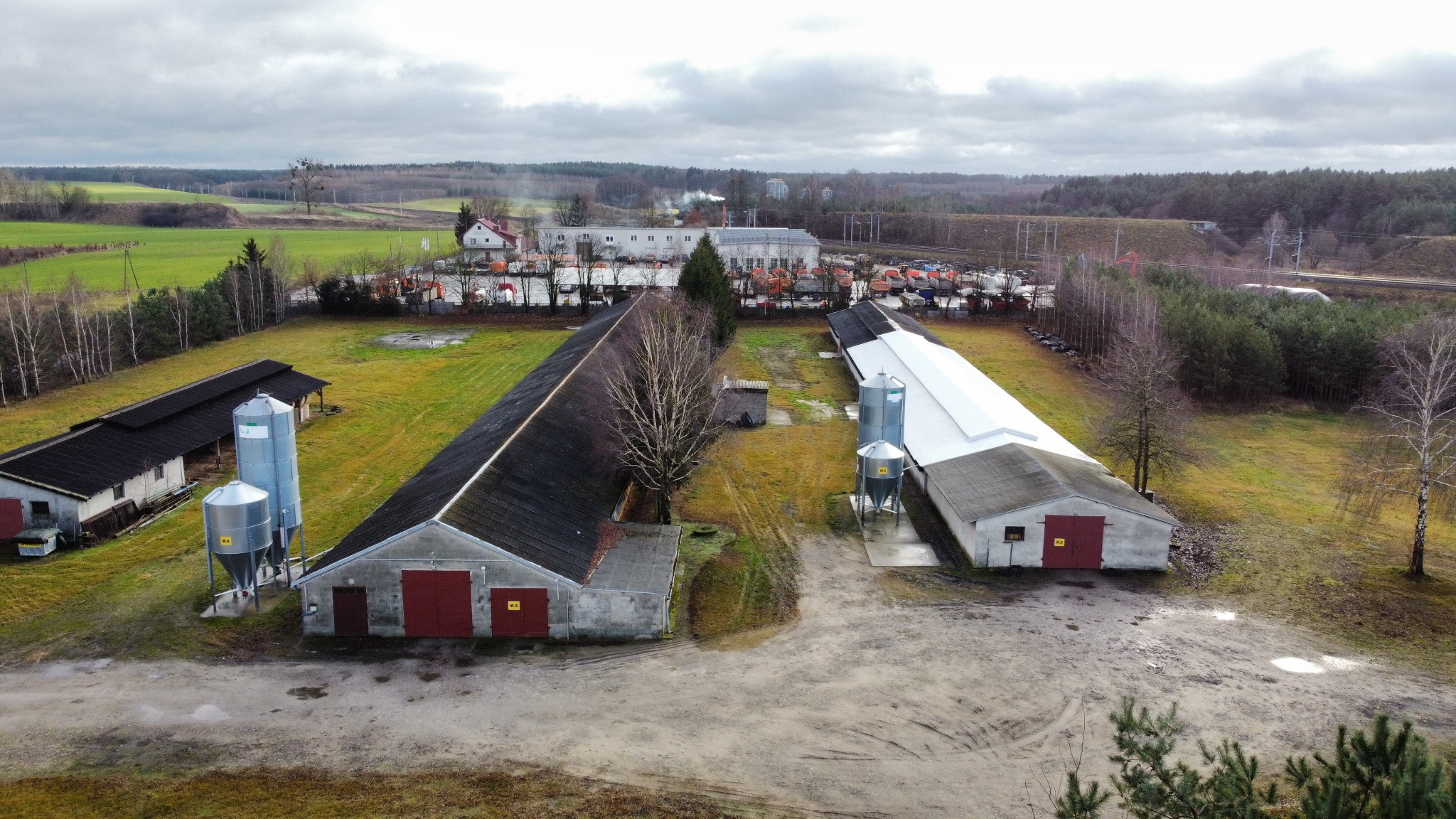 Widok z lotu ptaka na kompleks budynków gospodarczych z ciemnymi dachami i czerwonymi bramami, uzupełnionych o metalowe silosy, na tle rozległych pól i lasów pod pochmurnym niebem.