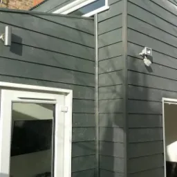 Elewacja domu w Gozdawie wykończona szarą, poziomą boazerią, z widocznymi białymi oknami i drzwiami oraz rynną.