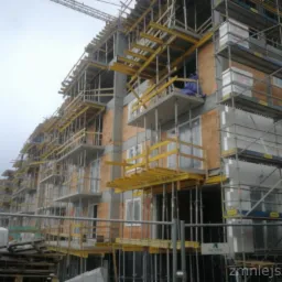 Wielopiętrowy budynek w trakcie budowy, z widocznymi elementami konstrukcyjnymi, rusztowaniami i częściowo ocieplonymi ścianami, sfotografowany w pochmurny dzień.