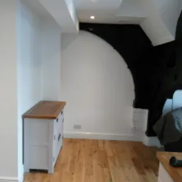 Jasne drewniane panele podłogowe w pomieszczeniu z białymi ścianami i komodami w stylu rustykalnym, widoczny fragment czarnego łuku i ozdobny element dekoracyjny ściany.