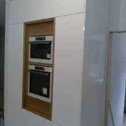 Biała, lakierowana zabudowa kuchenna z dwoma piekarnikami w drewnianej obudowie, widoczna z boku, z elementami wykończeniowymi w tle.