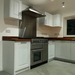 Jasna kuchnia z białymi szafkami i drewnianym blatem, duży stalowy piekarnik z okapem, szare płytki podłogowe, widoczne gniazdka elektryczne na ścianie.