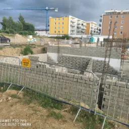 Wznoszone ściany parteru domu z betonu komórkowego na fundamencie, zabezpieczone barierkami z żółtą tablicą ostrzegawczą, na tle budynków mieszkalnych i żurawia budowlanego.